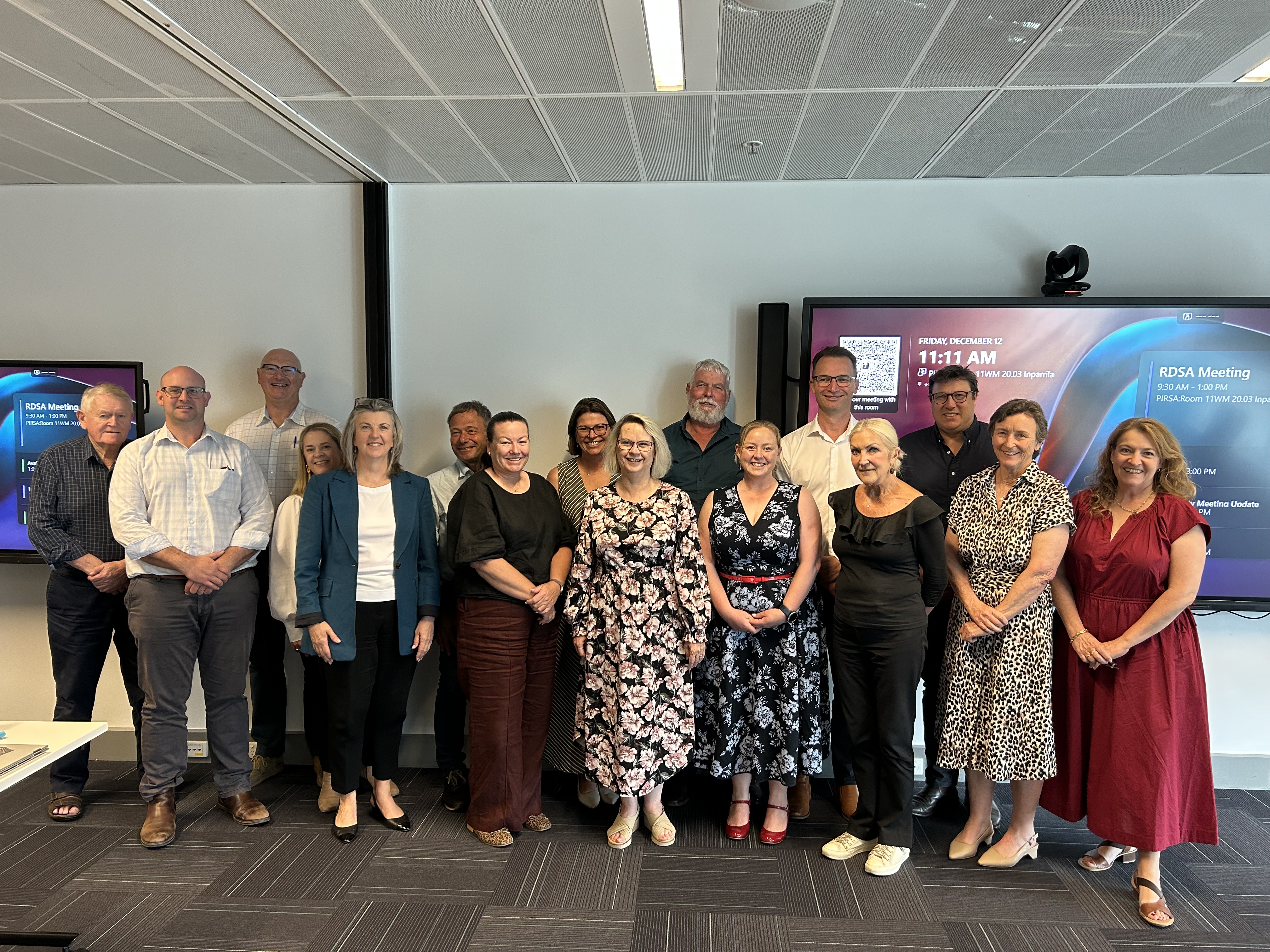 Group of sixteen adults standing in a meeting room with two digital screens displaying RDSA meeting details in the background.