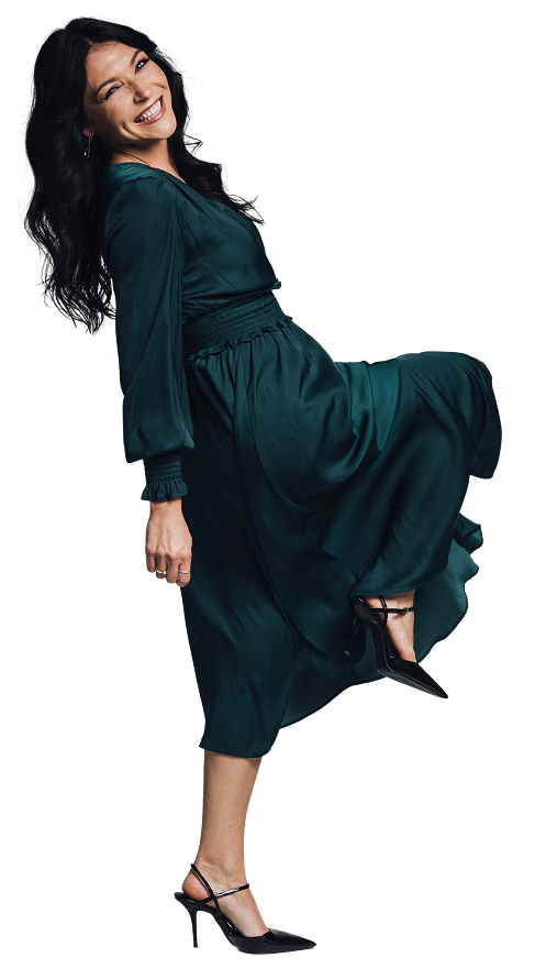 Smiling woman in a flowing dark green dress and black heels lifting one leg playfully.