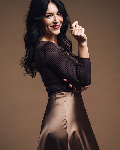 Woman with long black hair wearing a dark ribbed top and a shiny brown skirt, posing confidently against a brown background.