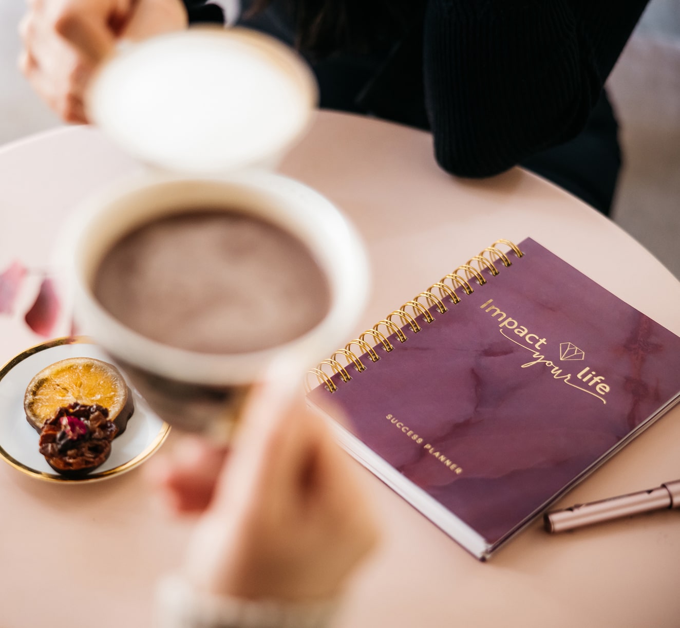 Close-up of a person holding a cup of coffee near a purple spiral-bound success planner and a small plate with dried fruits on a round table.
