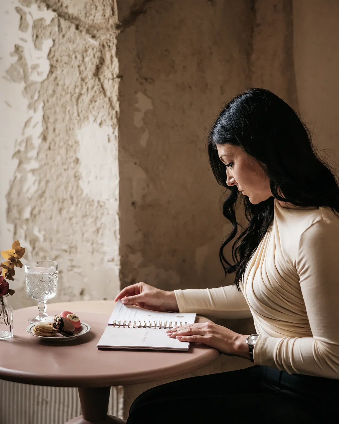 Woman with long black hair reading a book at a round table with a glass of water and macarons on a plate, against a rustic textured wall.
