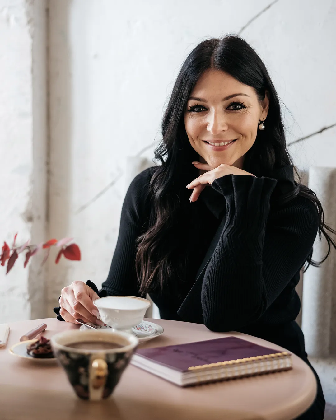 Smiling woman with long dark hair in a black sweater holding a white teacup at a table with a notebook and a cup of coffee.