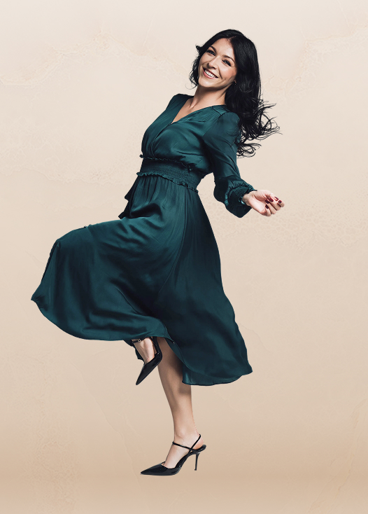 Smiling woman with long dark hair in a flowing dark green dress and black heels, captured mid-spin against a beige background.