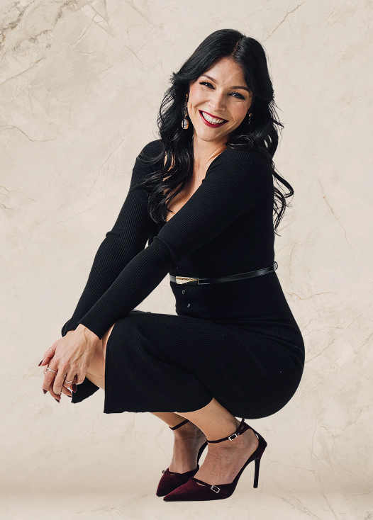 Woman with long black hair wearing a fitted black dress and burgundy high heels squatting and smiling against a light marble background.
