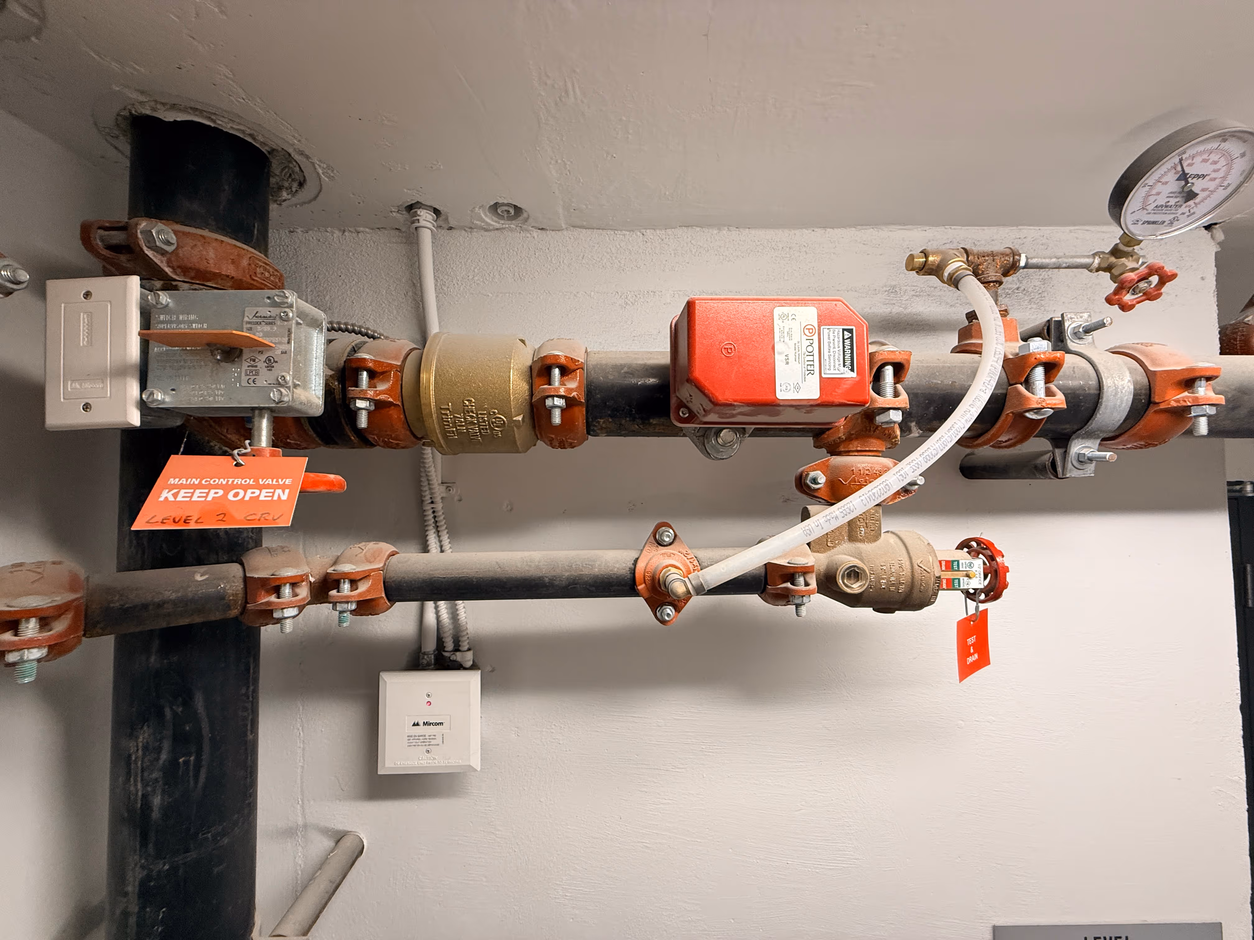 Industrial pipes with valves, pressure gauge, and red tags reading 'KEEP OPEN' and 'TEST & DRAIN.'