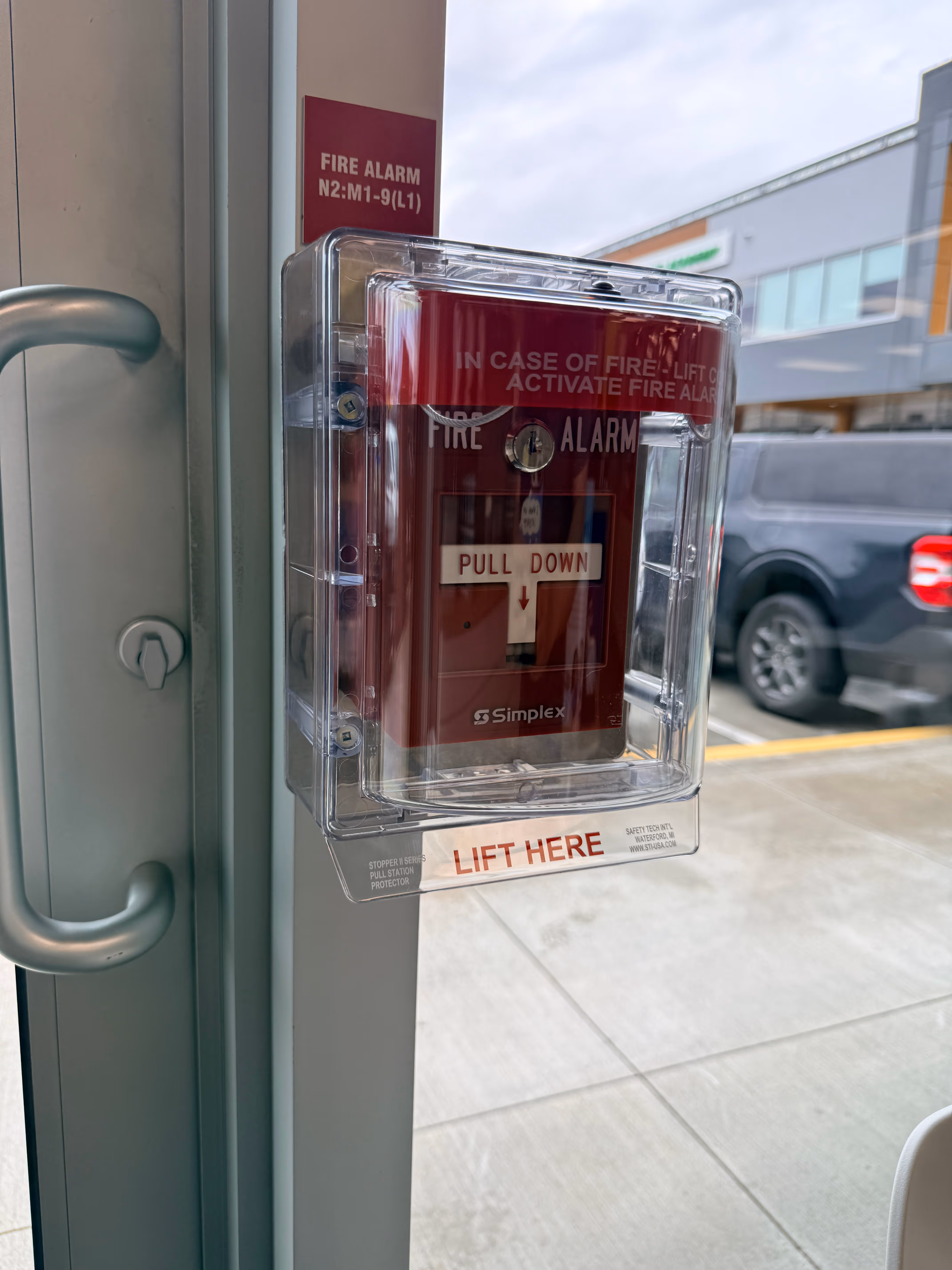 Red fire alarm pull station protected by clear plastic cover with 'Lift Here' instructions.