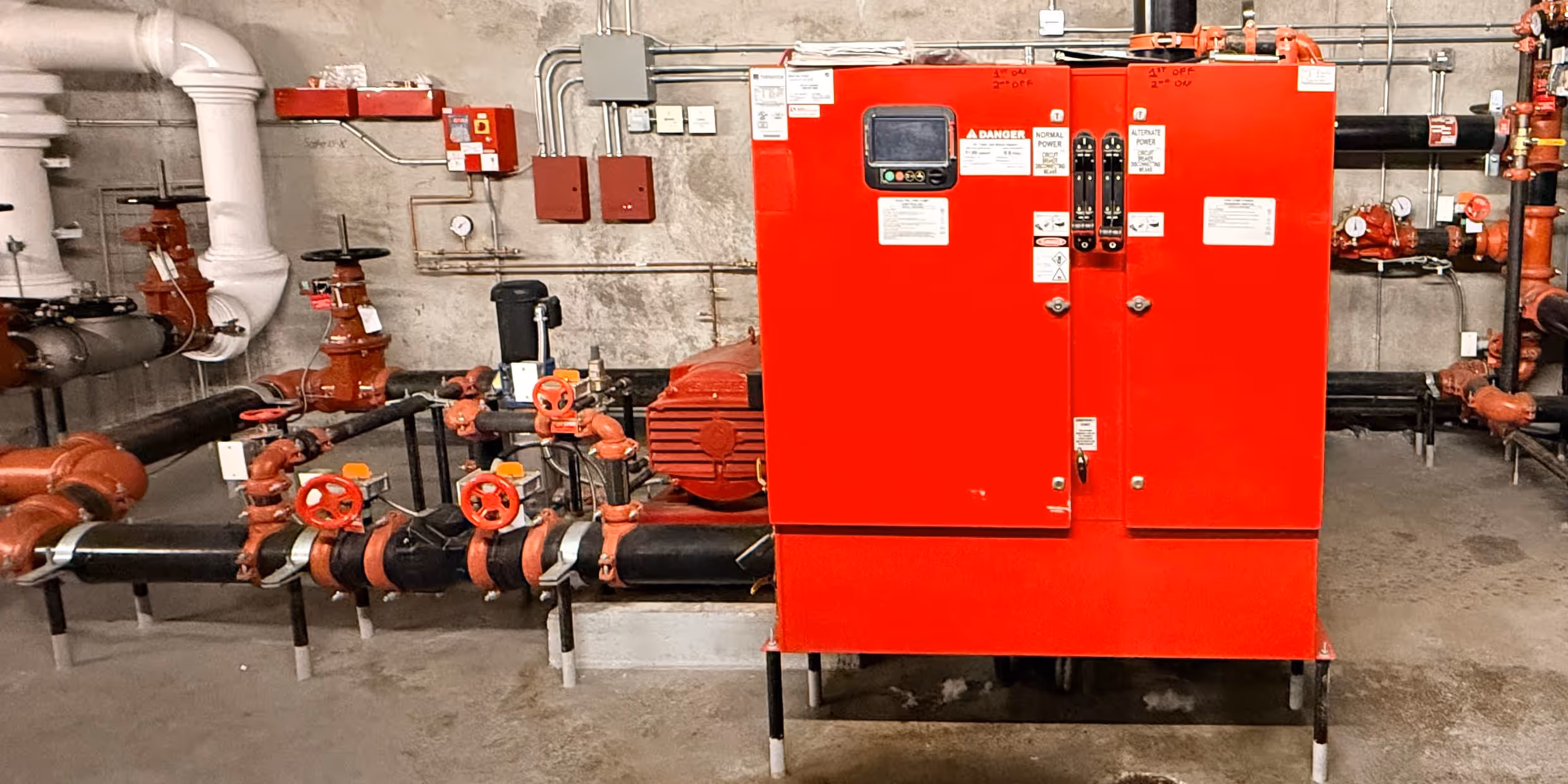 Industrial red fire pump control panel with black and red pipes and valves in a concrete room.