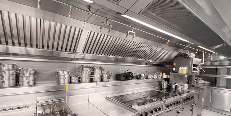 Commercial kitchen with stacked pots, stainless steel stove, industrial ventilation hood, and fryer baskets.
