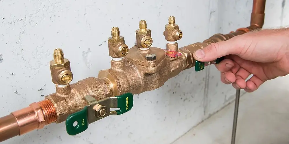 Hand adjusting a valve on a brass plumbing backflow preventer attached to copper pipes on a wall.