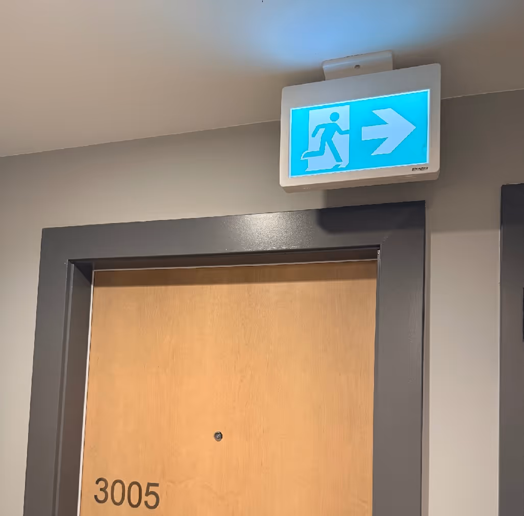 Wooden door numbered 3005 with an illuminated emergency exit sign above pointing right.