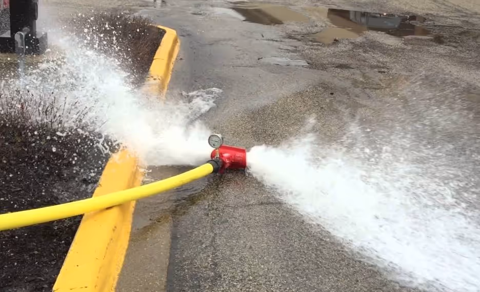 Yellow hose attached to a red spray nozzle releasing high-pressure water on pavement near a yellow curb.
