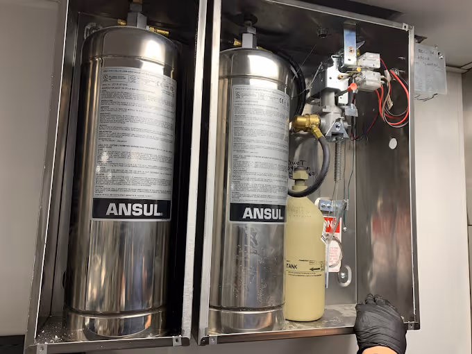 Two stainless steel Ansul fire suppression cylinders installed in a metal cabinet with a hand nearby.