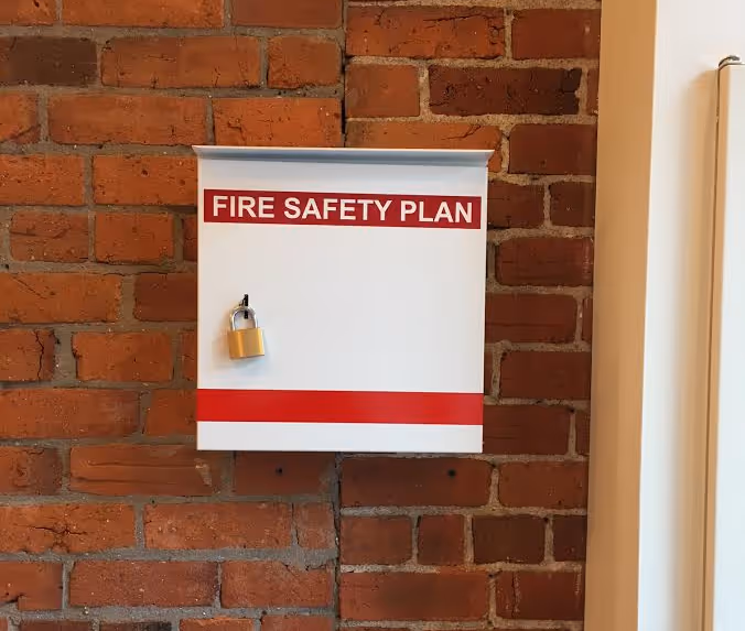 White metal box labeled "FIRE SAFETY PLAN" with red stripes and a padlock on a brick wall.