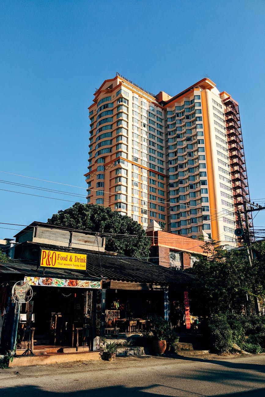 A modern skyscraper towers over a local diner in Chiang Mai, Thailand, under a clear blue sky.