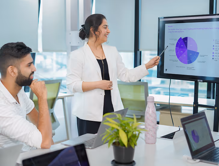 Woman in white blazer giving a presentation pointing at a pie chart on a screen while colleagues listen in a modern office.