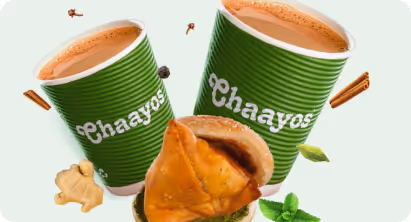 Two green paper cups of Chaayos chai with a samosa and floating spices like ginger, cardamom, and cinnamon sticks.