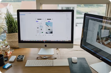 Office desk with two computer monitors displaying design and email applications, keyboard, mouse, glasses, and office supplies.
