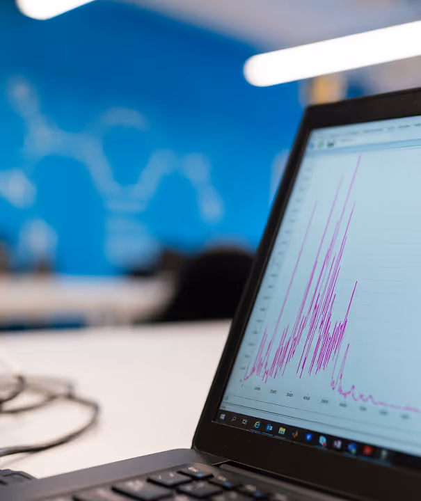 Laptop screen displaying a graph with purple spikes and trends, set in an office environment with a blurred blue background.