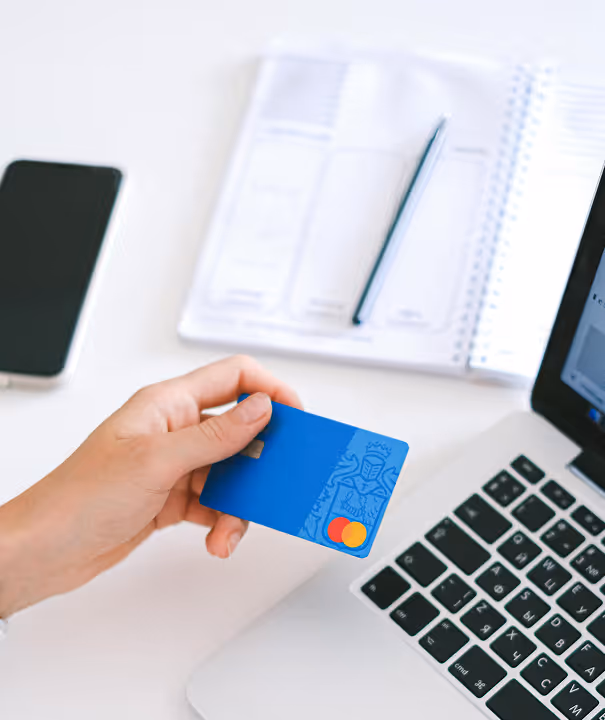 Hand holding a blue Mastercard credit card near a laptop keyboard, with a phone and notebook in the background.