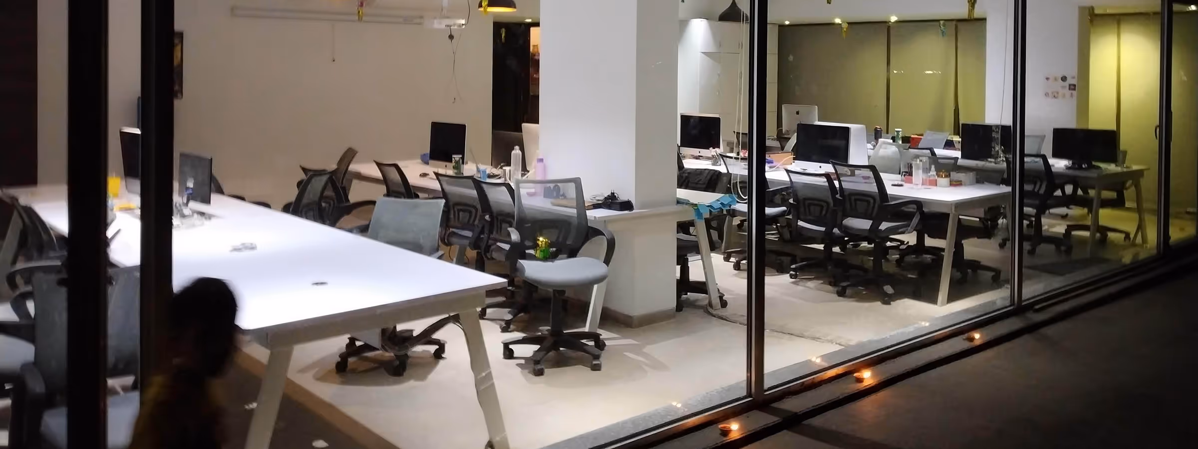 View through glass walls of a modern office with white desks, black swivel chairs, and computers at night.