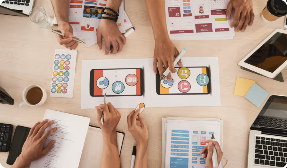 Overhead view of four people collaborating on a UX design project at a desk with smartphones, design layouts, markers, and laptops.