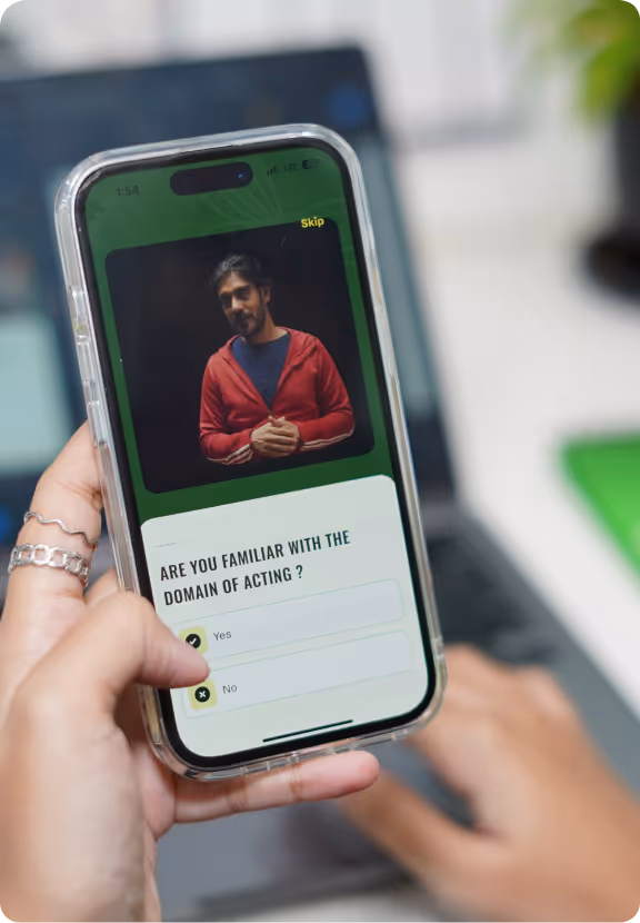 Hand holding a smartphone showing a man in a red jacket on a video call with a poll asking 'Are you familiar with the domain of acting?' and options Yes and No.