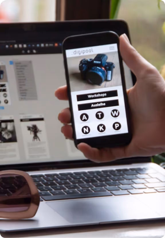 Hand holding smartphone displaying a camera rental and workshop website in front of a laptop with a similar webpage open.