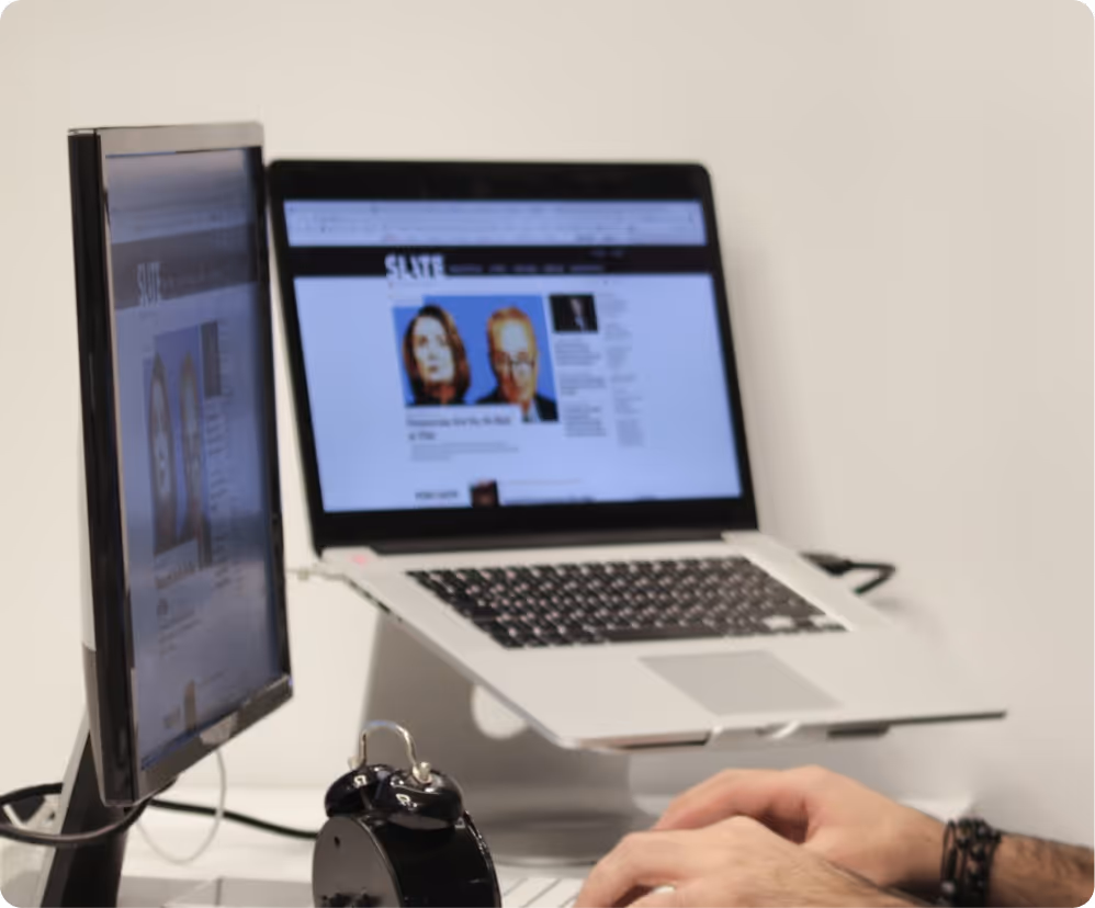 Person typing on a laptop at a desk with an additional monitor and a black alarm clock nearby.
