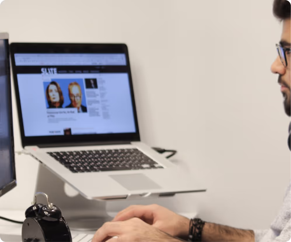 Person typing on a keyboard next to an alarm clock with a laptop displaying a website on a stand.