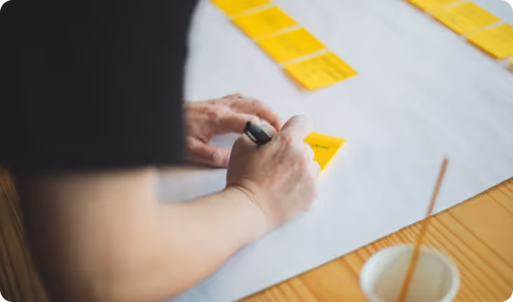 Person writing on yellow sticky notes arranged on a white surface over a wooden table.