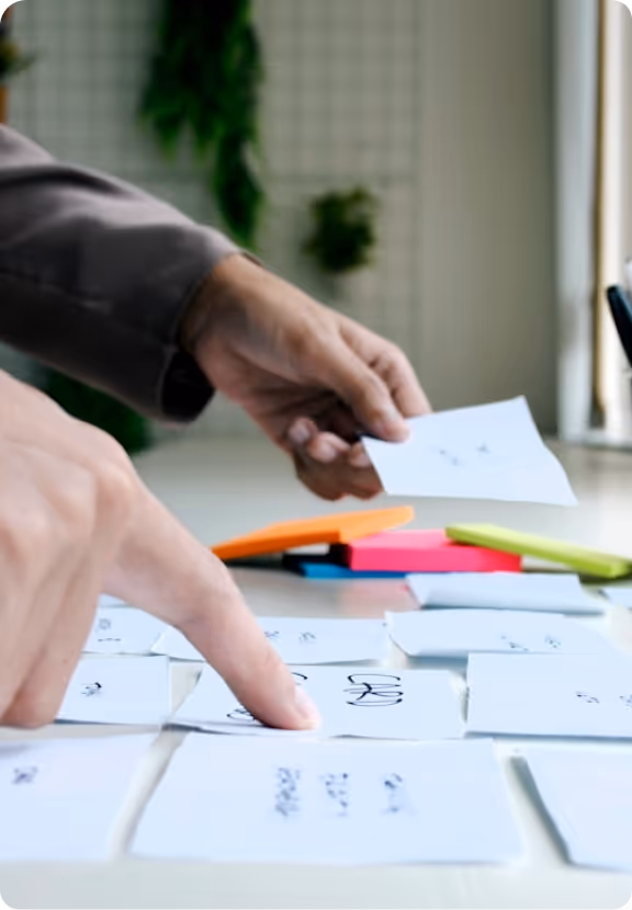Two hands organizing white index cards with handwritten notes on a table with colorful sticky notes in the background.