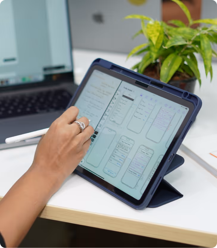 Person using a stylus to draw mobile app wireframes on a tablet, with a laptop and a plant in the background.