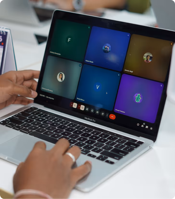 Hands typing on a MacBook Pro during a virtual video meeting with six participants displayed on the screen.