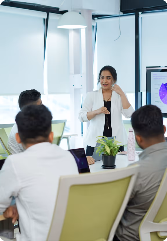 Woman in white blazer presenting to colleagues in a modern office meeting room.