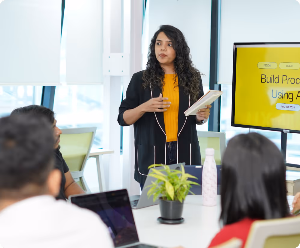 Woman giving a presentation to colleagues in a modern office meeting room with a screen displaying 'Build Products Using AI' and a table with laptops and a plant.