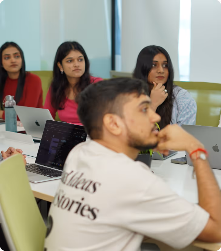 Group of young professionals seated around a conference table, engaged and listening during a meeting with laptops open.