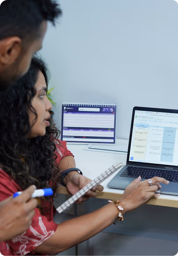 Two people discussing work while looking at a laptop screen displaying a spreadsheet and holding a notebook and marker.