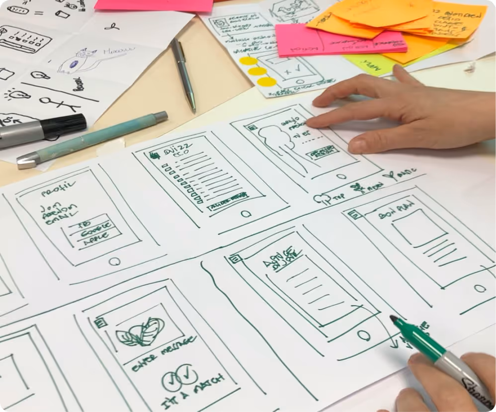 Person's hands sketching mobile app wireframes on paper surrounded by pens and colorful sticky notes.