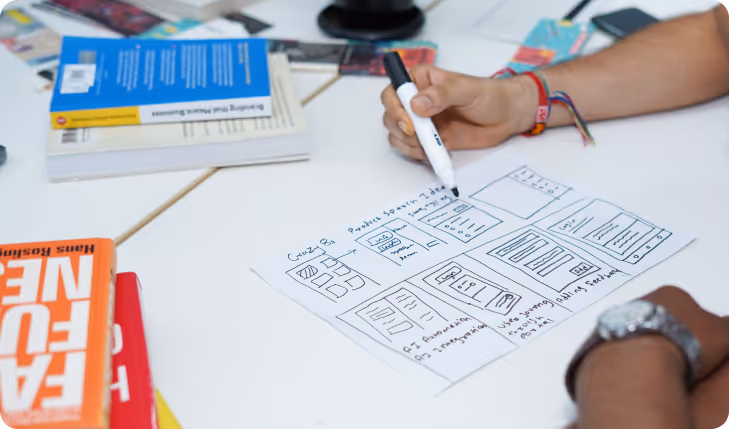 Person sketching a website wireframe on paper with a black marker, with colorful books on a white table.