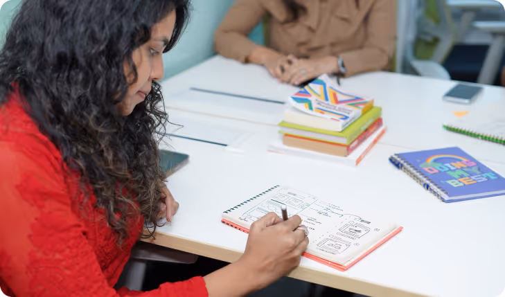 Woman in red shirt writing sketches and notes in a notebook at a white table with another person and stacked books.