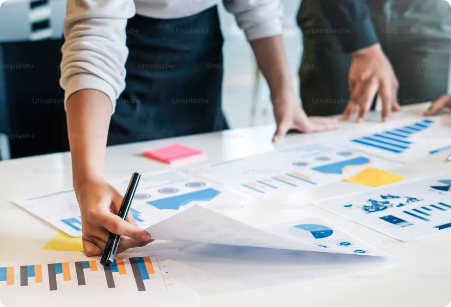 Two people reviewing printed charts and graphs on a table, with one person holding a pen and pointing at a bar graph.