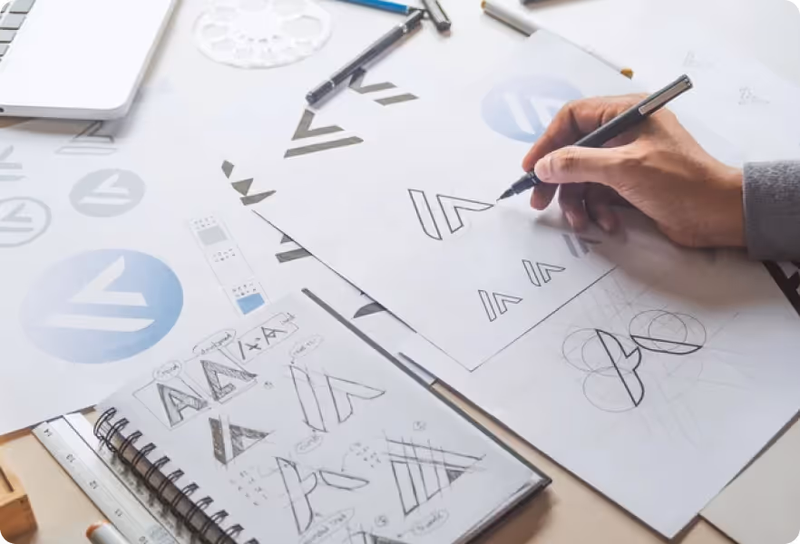 Hand sketching minimalist geometric logo designs on paper surrounded by other logo drafts and drawing tools on a desk.