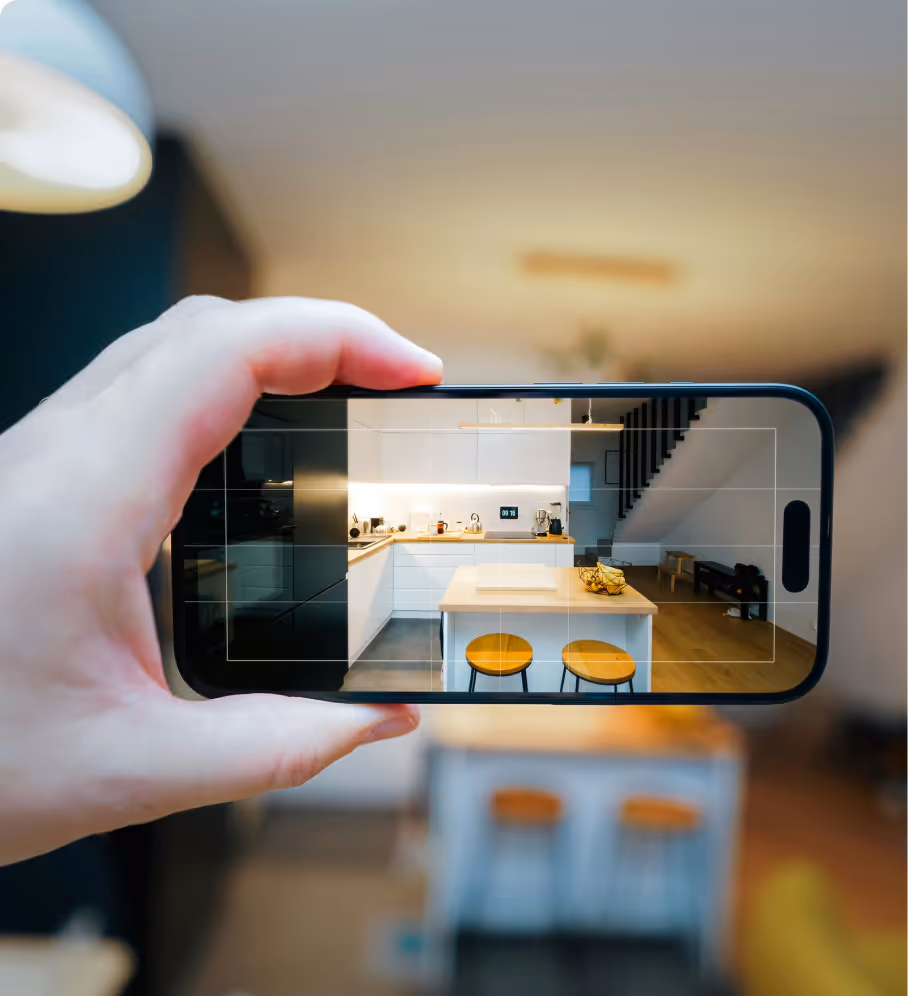 A hand holding a smartphone capturing a modern kitchen with white cabinets, wooden countertop, two stools, and a staircase in the background.