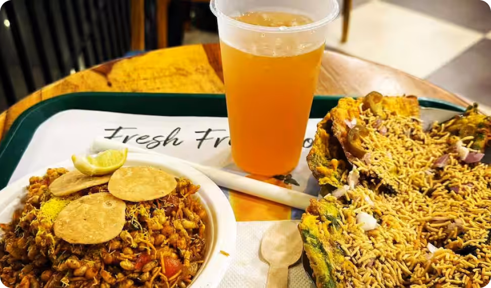 Traditional Indian snacks including a bowl of chana chaat with papdis and a lemon wedge, a plastic cup of orange juice, and a stuffed, fried snack topped with sev on a tray.