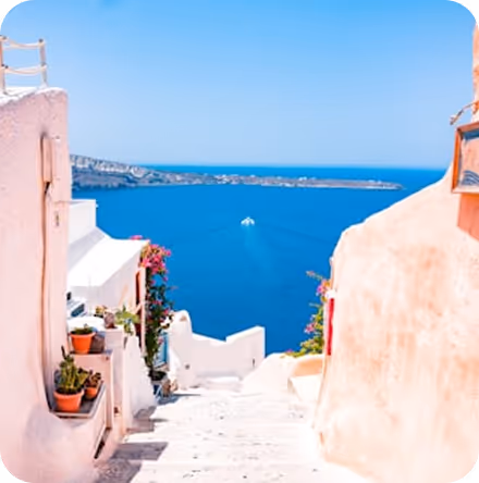 Stairway lined with white buildings and potted plants leading down to a blue sea with a boat in the distance under clear sky.