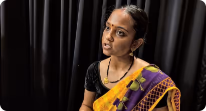 Woman wearing a yellow and purple sari speaking in front of black curtains.