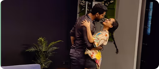 Couple embracing indoors near a green plant and smiling at each other.