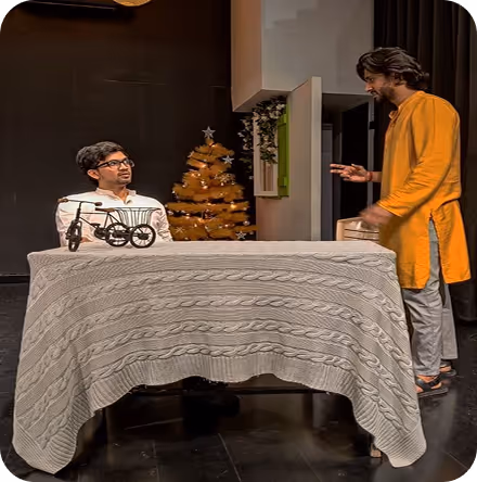Two men in a room with a decorated Christmas tree in the background; one man sits behind a table with a bicycle placed on it while the other stands gesturing.