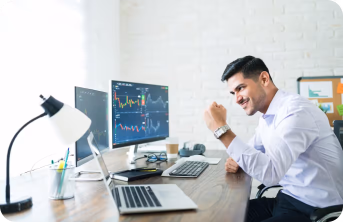 Smiling man in white shirt sitting at desk with multiple monitors showing stock market charts.