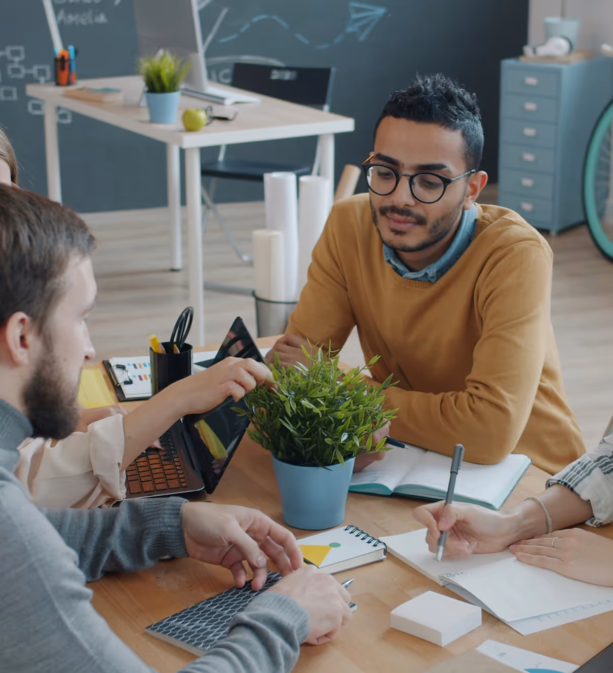 Group of people collaborating at a table with notebooks, a tablet, and a potted plant in an office setting.